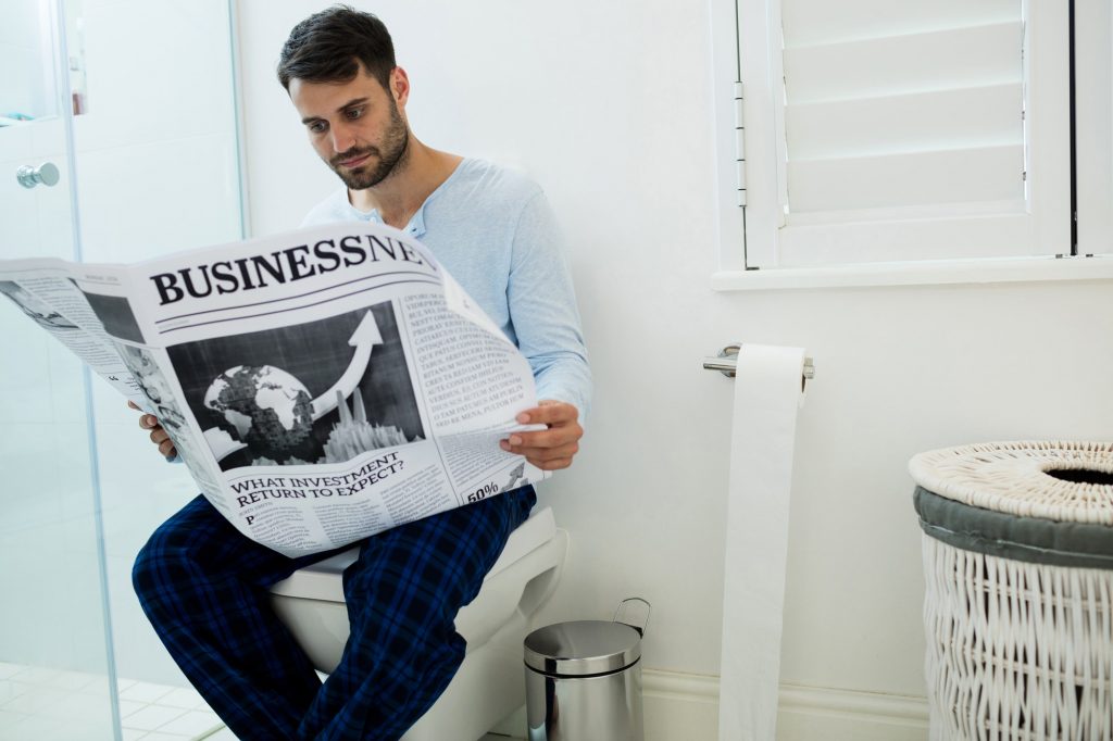 Man sitting on toilet seat reading a newspaper