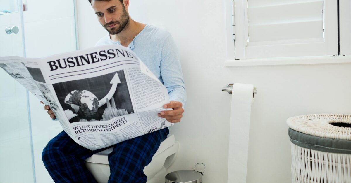 Man sitting on toilet seat reading a newspaper